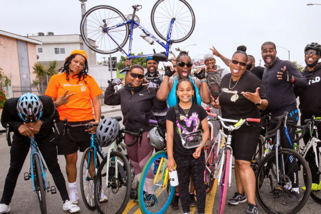 Eleven people of color, of all ages, pose with their bicycles, laughing and smiling together.